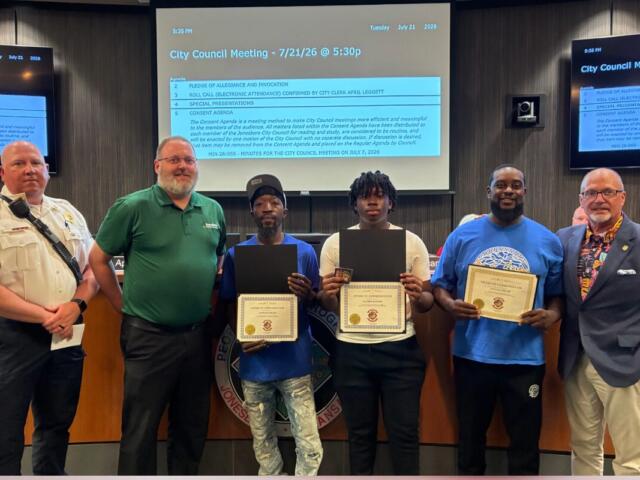 Six people stand behind a council desk; four award recipients hold certificates, flanked by officials during City Council meeting.
