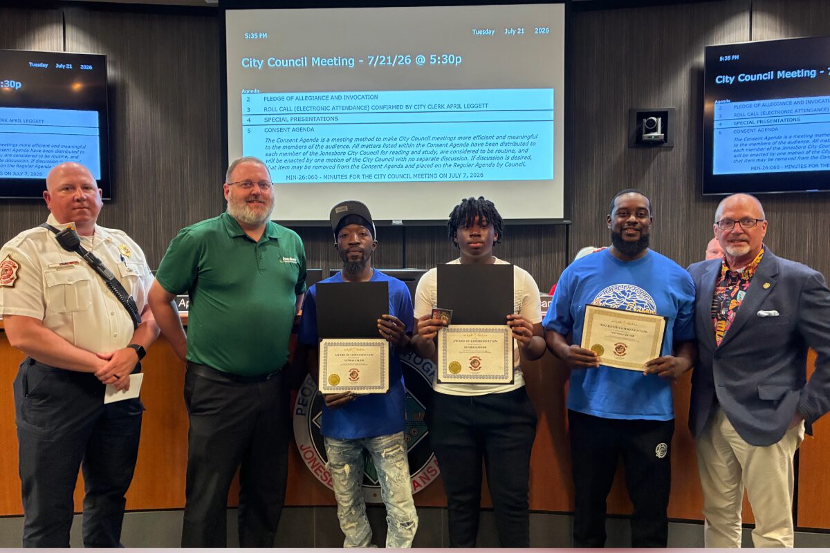 Six people stand behind a council desk; four award recipients hold certificates, flanked by officials during City Council meeting.