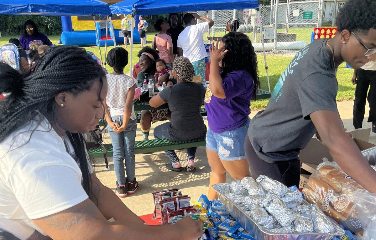 People gather under blue canopy tents at an outdoor event, serving and sharing snacks at a picnic tables setup.