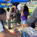People gather under blue canopy tents at an outdoor event, serving and sharing snacks at a picnic tables setup.