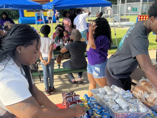 People gather under blue canopy tents at an outdoor event, serving and sharing snacks at a picnic tables setup.