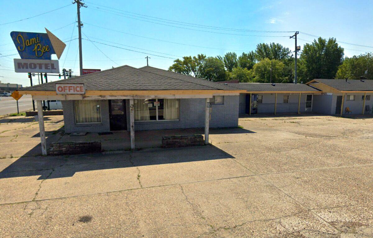 Low, single-story motel office with a faded 'Office' sign and a large roadside 'Dami Bee Motel' sign on the left. The parking area in front is cracked and empty.