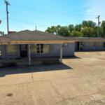 Low, single-story motel office with a faded 'Office' sign and a large roadside 'Dami Bee Motel' sign on the left. The parking area in front is cracked and empty.
