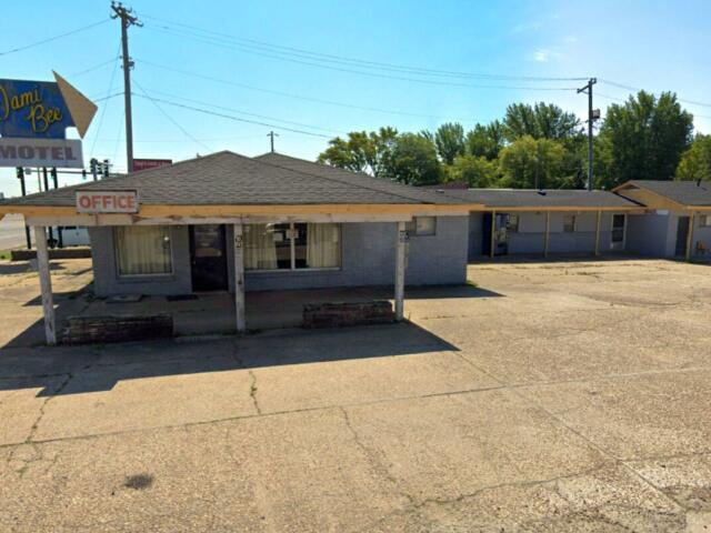 Low, single-story motel office with a faded 'Office' sign and a large roadside 'Dami Bee Motel' sign on the left. The parking area in front is cracked and empty.