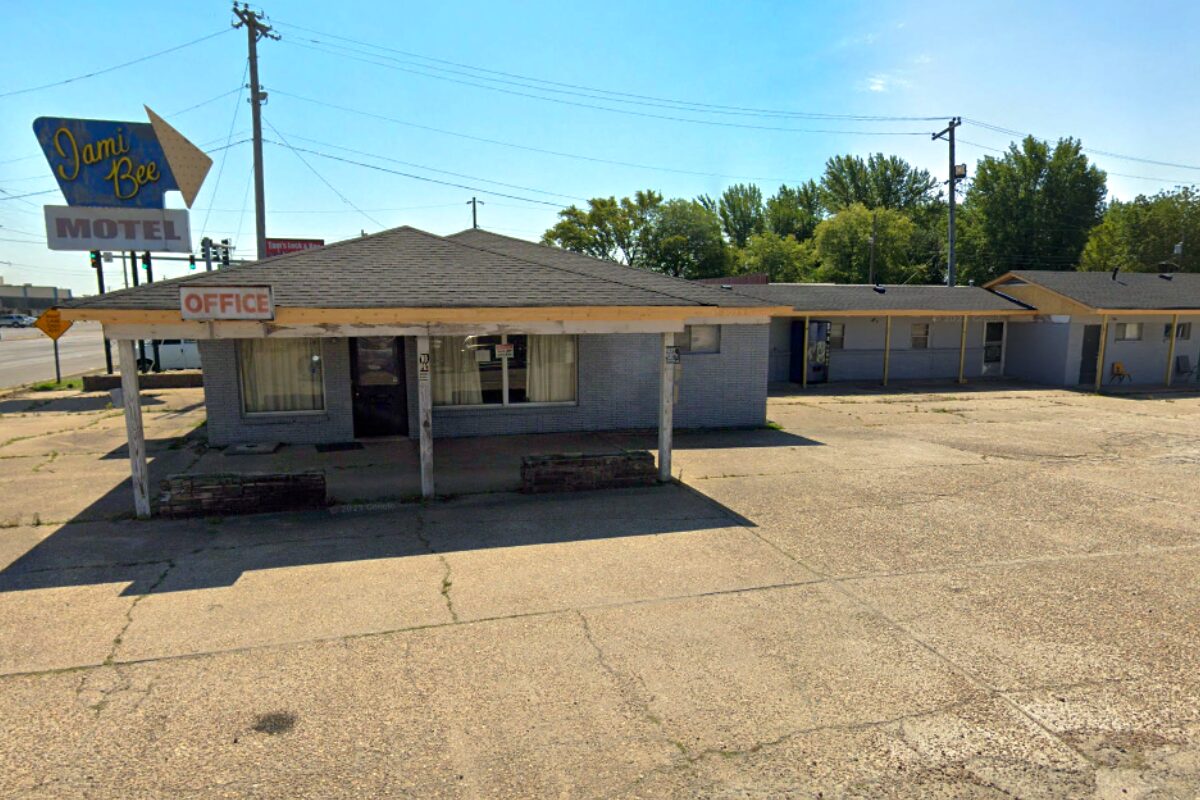 Low, single-story motel office with a faded 'Office' sign and a large roadside 'Dami Bee Motel' sign on the left. The parking area in front is cracked and empty.