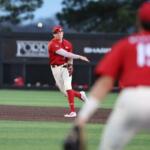 Baseball pitcher in red uniform throws a pitch on the mound, with a fielder in foreground wearing number 15 blurred in the foreground.