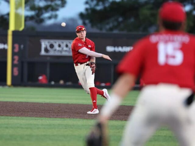 Baseball pitcher in red uniform throws a pitch on the mound, with a fielder in foreground wearing number 15 blurred in the foreground.