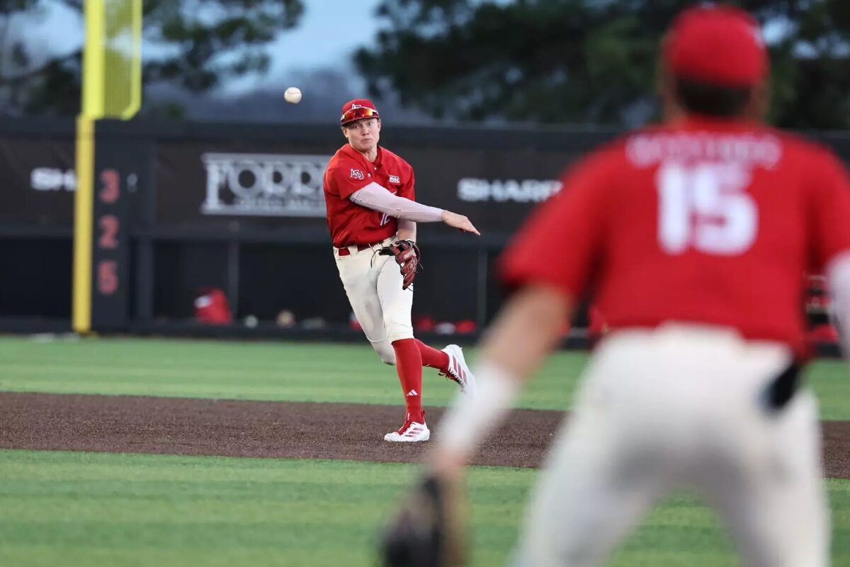Baseball pitcher in red uniform throws a pitch on the mound, with a fielder in foreground wearing number 15 blurred in the foreground.