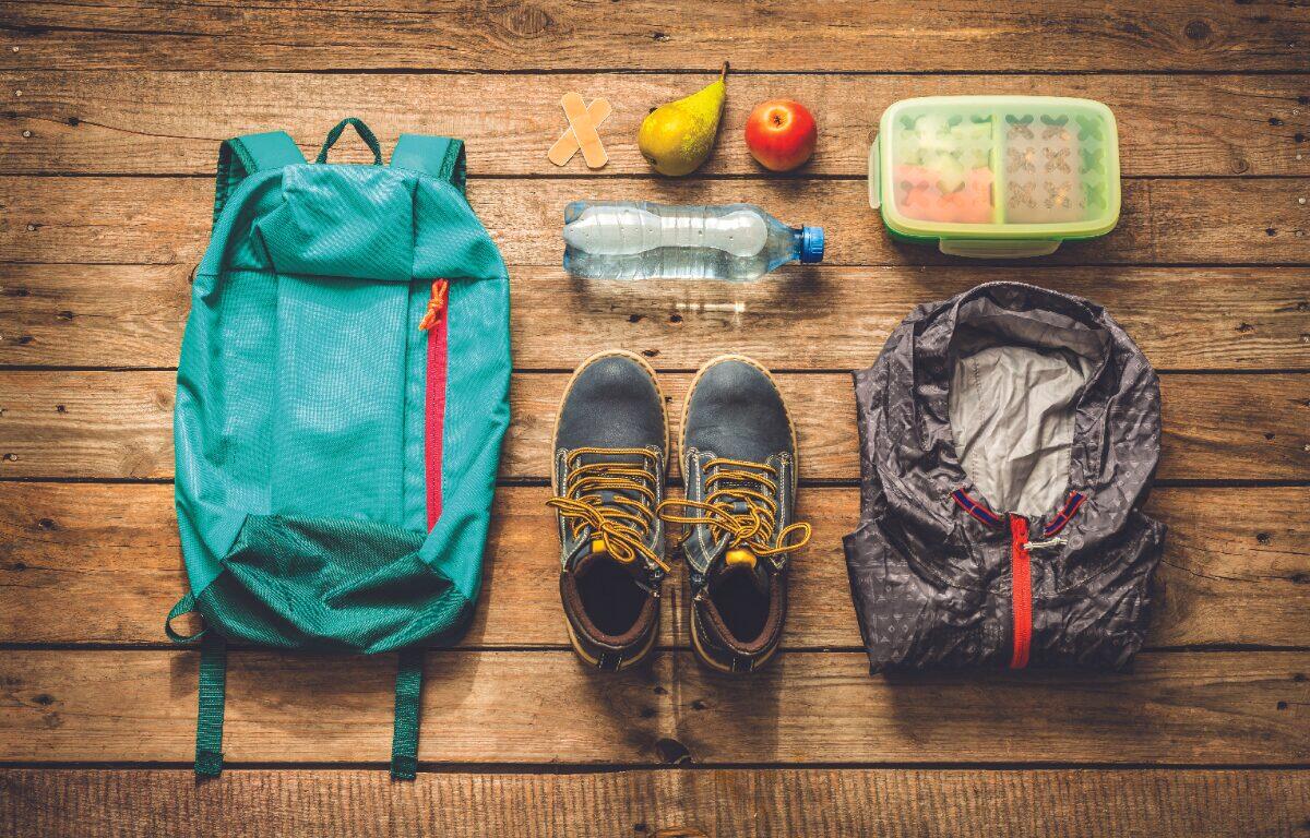 Hiking gear laid out on wooden planks: teal backpack, waterproof jacket, hiking boots, water bottle, fruit, lunchbox, and band-aids.