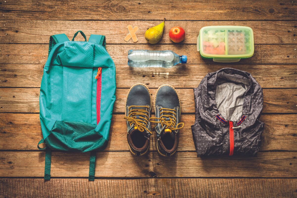 Hiking gear laid out on wooden planks: teal backpack, waterproof jacket, hiking boots, water bottle, fruit, lunchbox, and band-aids.