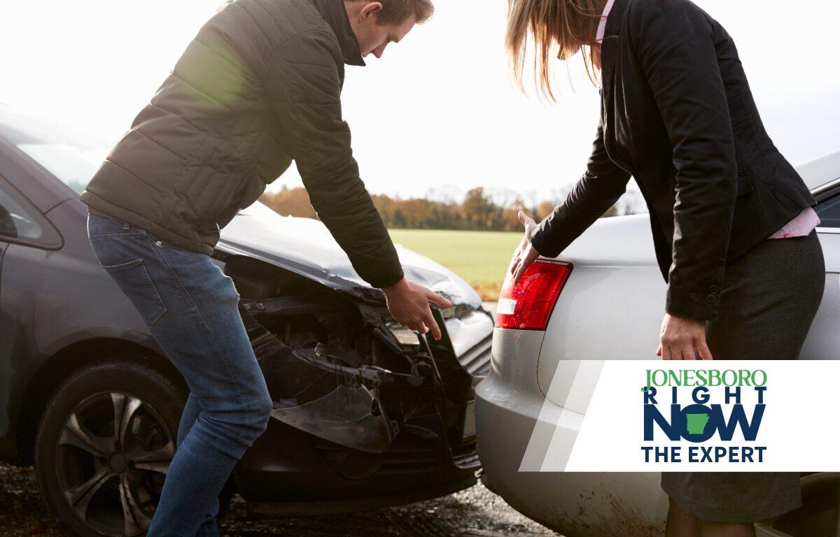Two people inspect the front ends of two cars after a collision, with a news logo overlay in the corner.