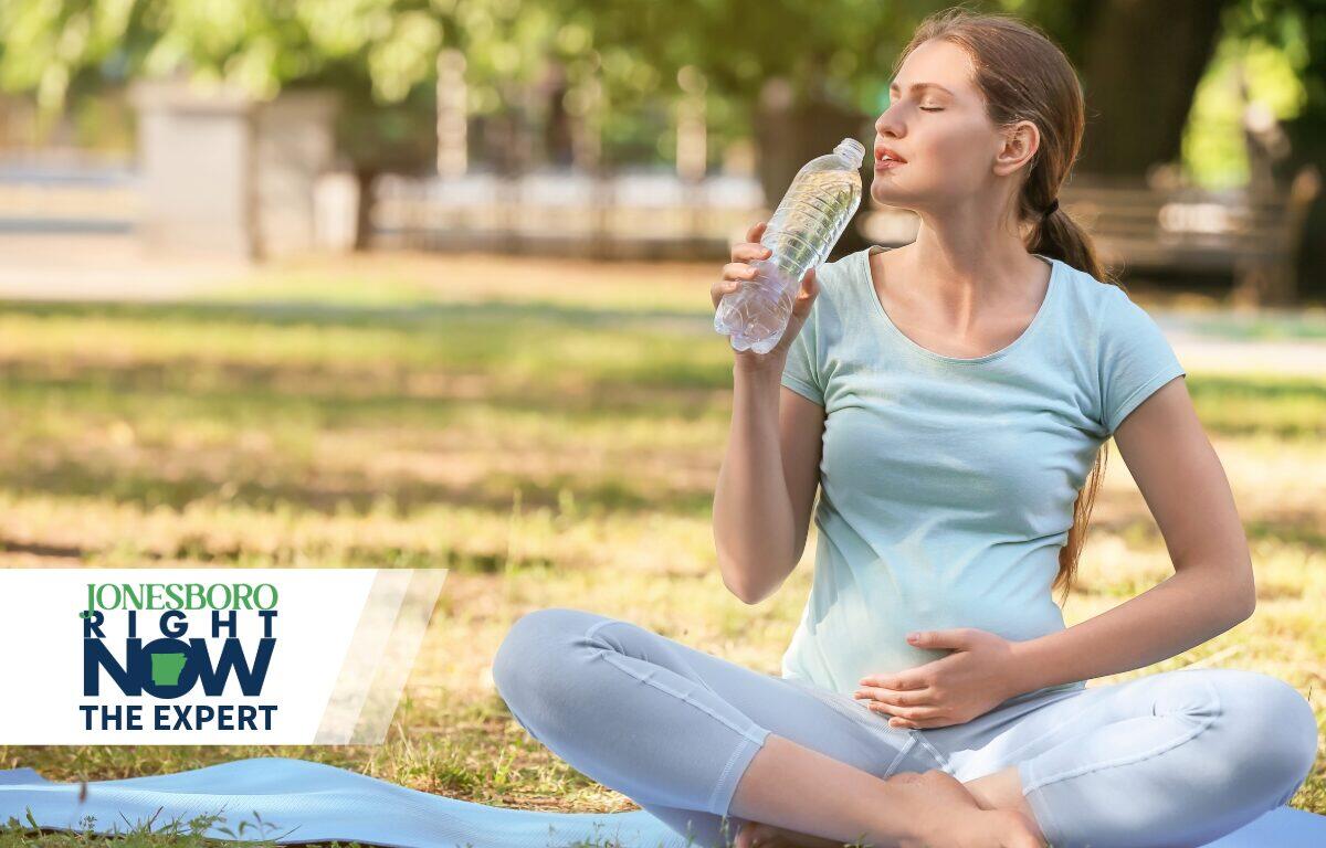 Pregnant or exercises woman sitting cross‑legged on a yoga mat in a park, sipping water with eyes closed; Jonesboro Right Now logo in the lower left corner.