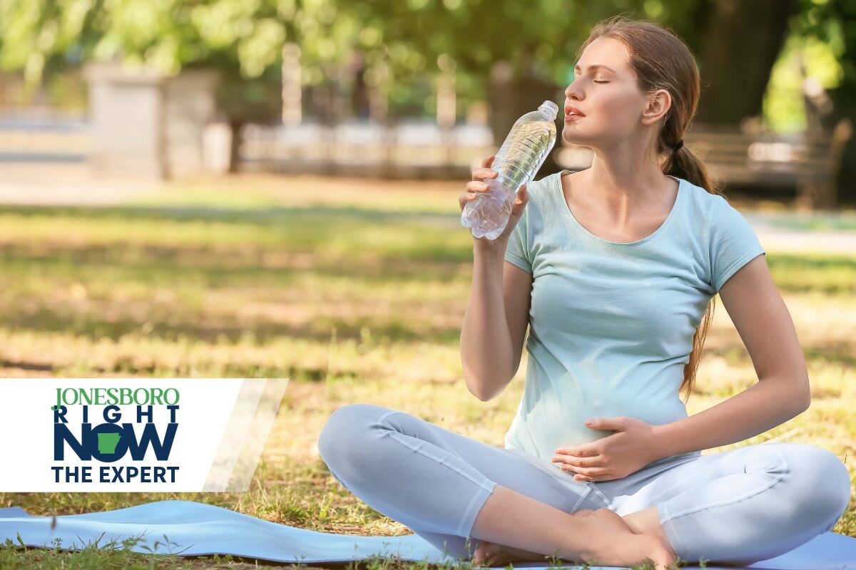 Pregnant or exercises woman sitting cross‑legged on a yoga mat in a park, sipping water with eyes closed; Jonesboro Right Now logo in the lower left corner.