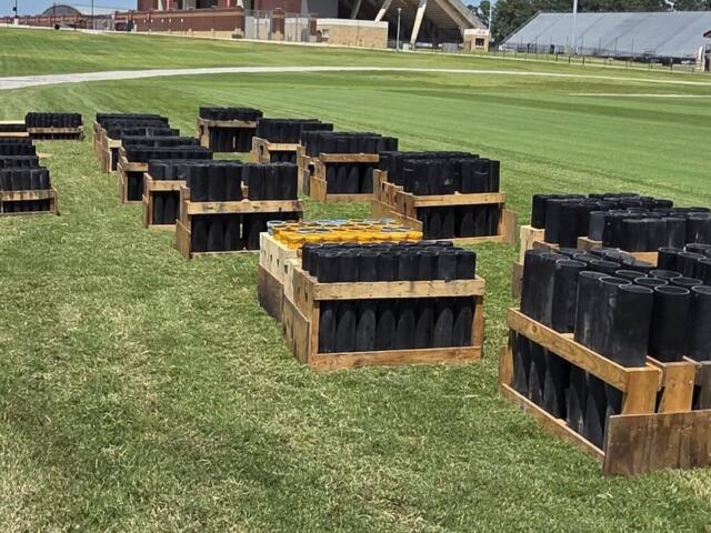 Pallets of black cylindrical tubes stacked on grass at a sports field, with a stadium in the background.