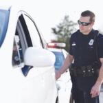 Police officer in a dark uniform checks a white car at a roadside stop, emergency lights in the background.
