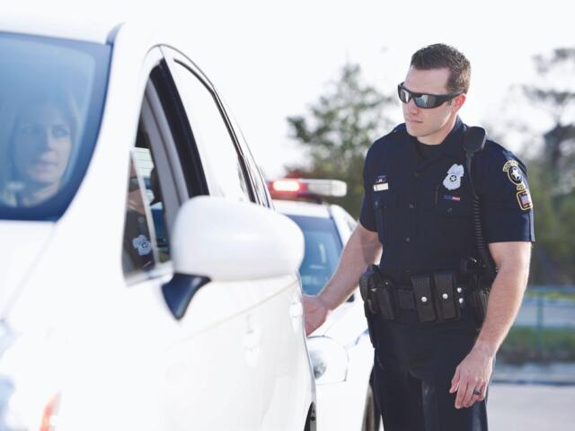 Police officer in a dark uniform checks a white car at a roadside stop, emergency lights in the background.