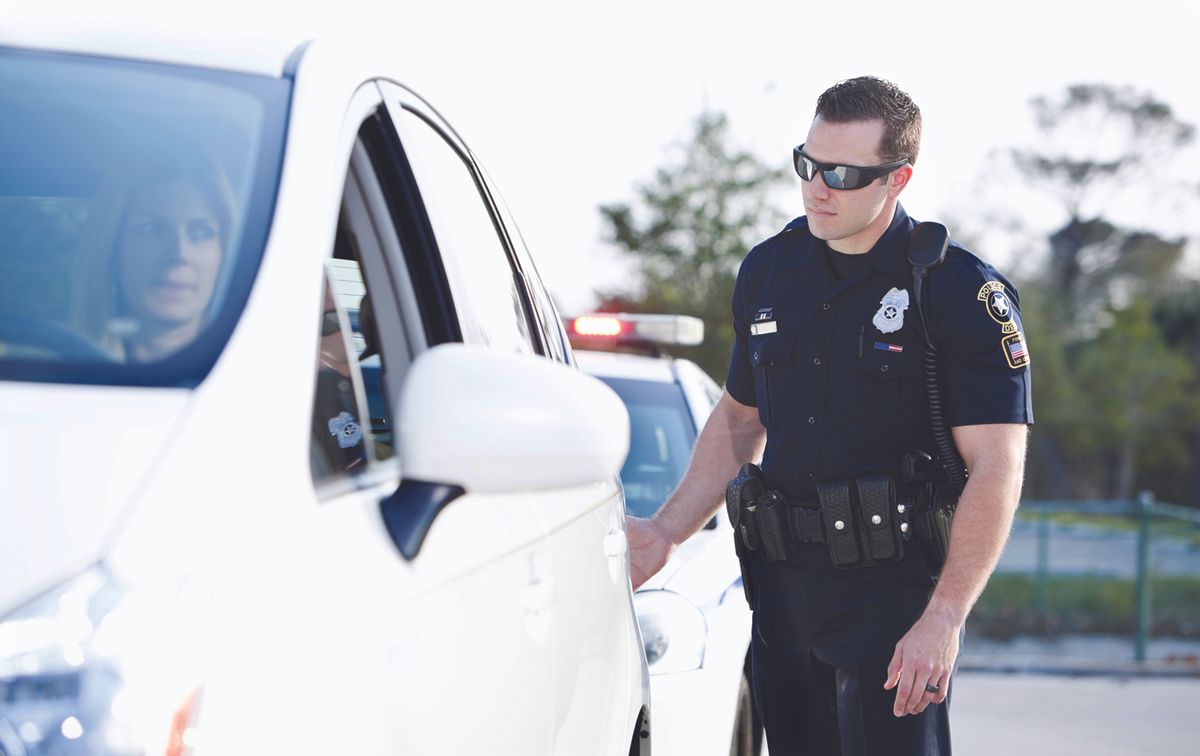 Police officer in a dark uniform checks a white car at a roadside stop, emergency lights in the background.