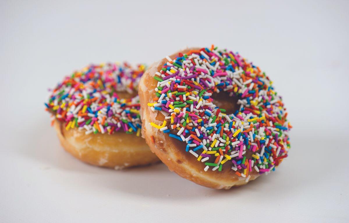 Two donuts with colorful rainbow sprinkles sit on a white surface, one foregrounded with chocolate glaze and sprinkles visible.