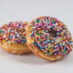 Two donuts with colorful rainbow sprinkles sit on a white surface, one foregrounded with chocolate glaze and sprinkles visible.