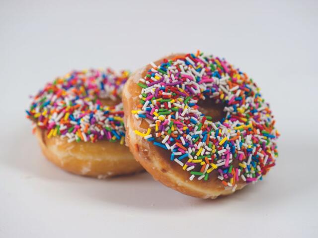 Two donuts with colorful rainbow sprinkles sit on a white surface, one foregrounded with chocolate glaze and sprinkles visible.