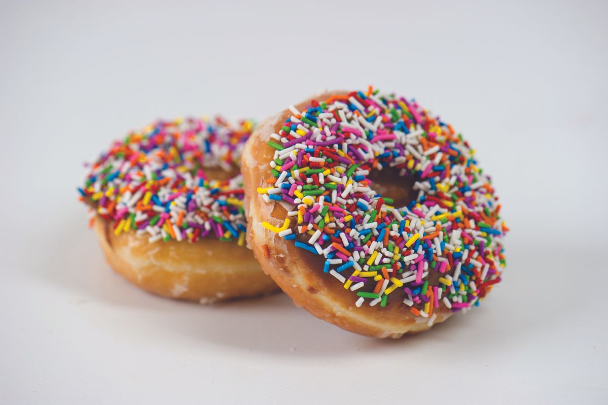 Two donuts with colorful rainbow sprinkles sit on a white surface, one foregrounded with chocolate glaze and sprinkles visible.