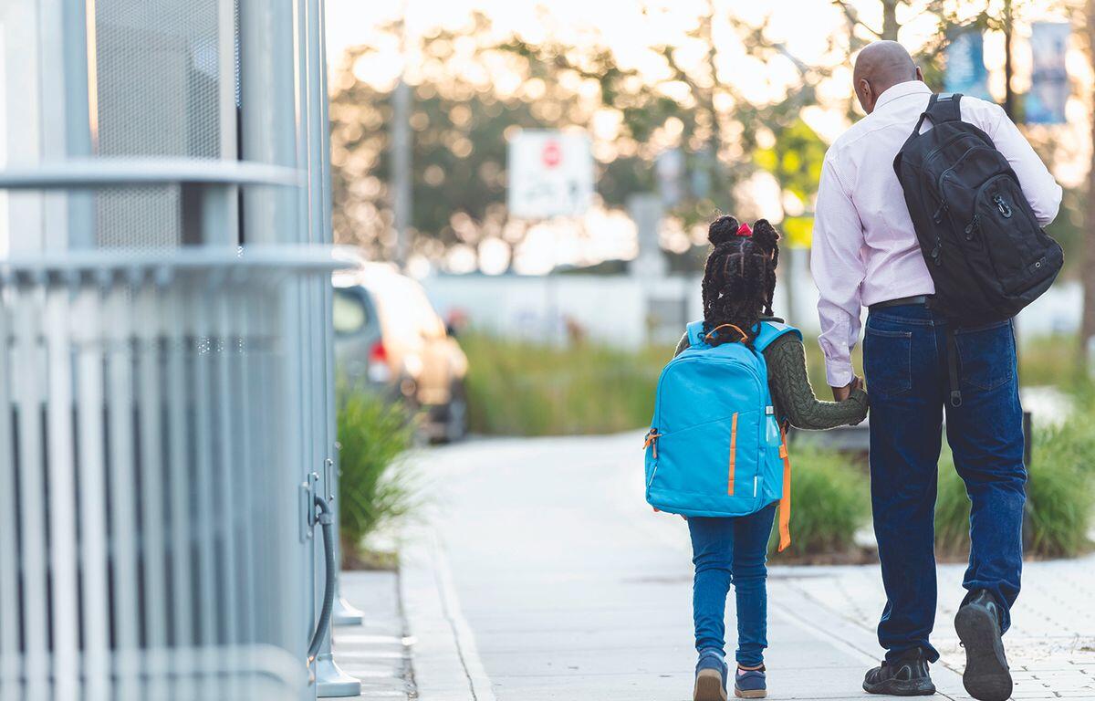 Man and child walk hand in hand on a sunny sidewalk, both wearing backpacks.