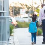 Man and child walk hand in hand on a sunny sidewalk, both wearing backpacks.