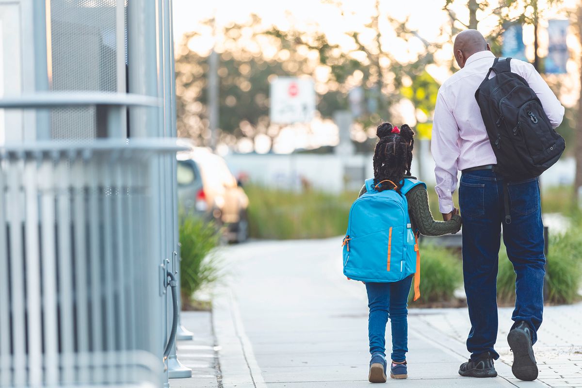 Man and child walk hand in hand on a sunny sidewalk, both wearing backpacks.