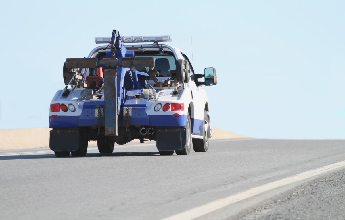 Blue-and-white tow truck with heavy towing gear and crane at the rear on a road, clear sky behind.