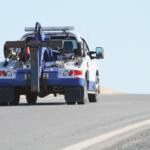 Blue-and-white tow truck with heavy towing gear and crane at the rear on a road, clear sky behind.