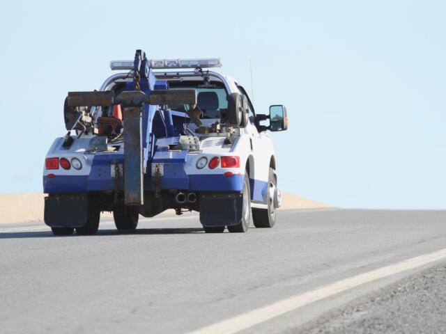 Blue-and-white tow truck with heavy towing gear and crane at the rear on a road, clear sky behind.