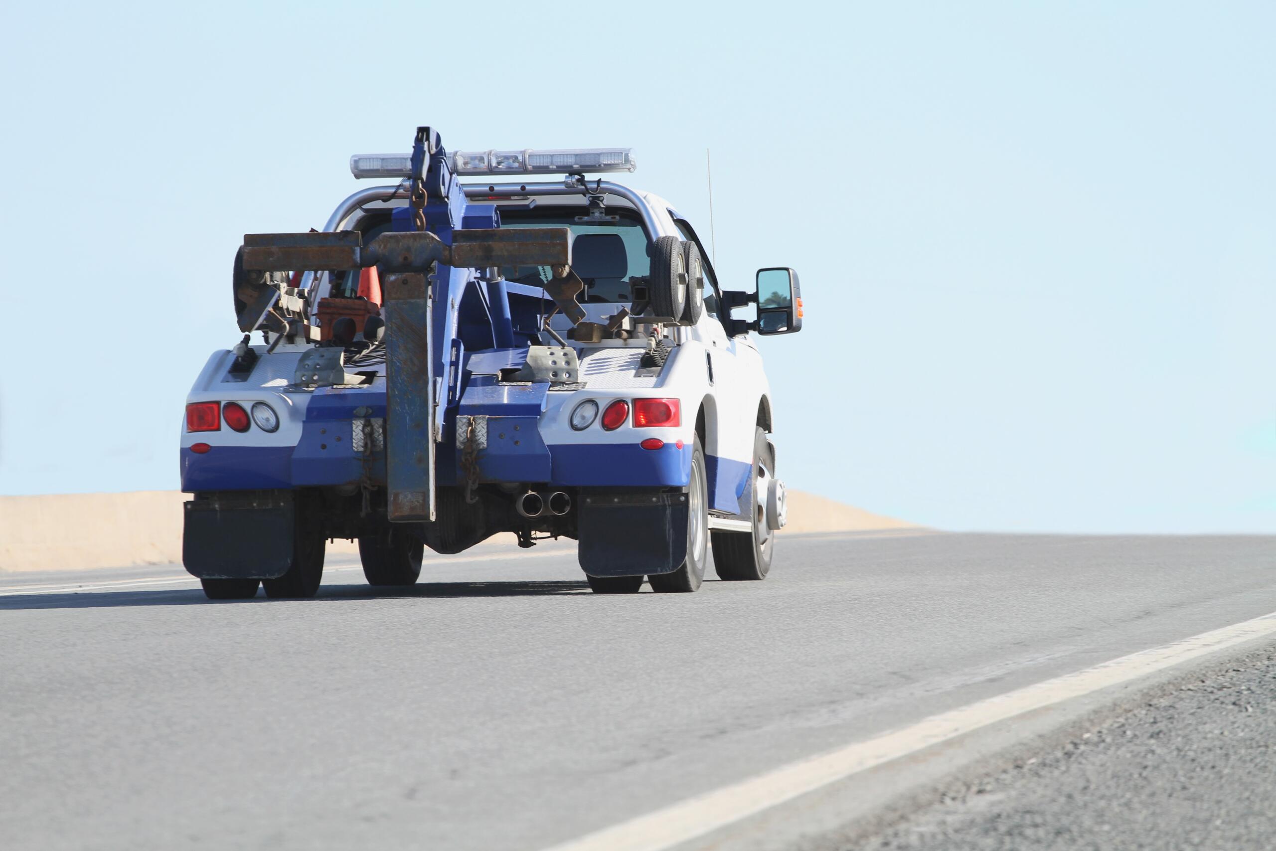 Blue-and-white tow truck with heavy towing gear and crane at the rear on a road, clear sky behind.