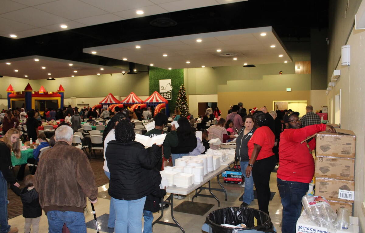 Crowded indoor event in a hall with long tables, plastic chairs, and inflatable red-and-yellow attractions in the background; a Christmas tree visible.