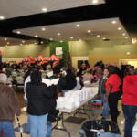 Crowded indoor event in a hall with long tables, plastic chairs, and inflatable red-and-yellow attractions in the background; a Christmas tree visible.