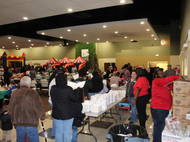 Crowded indoor event in a hall with long tables, plastic chairs, and inflatable red-and-yellow attractions in the background; a Christmas tree visible.