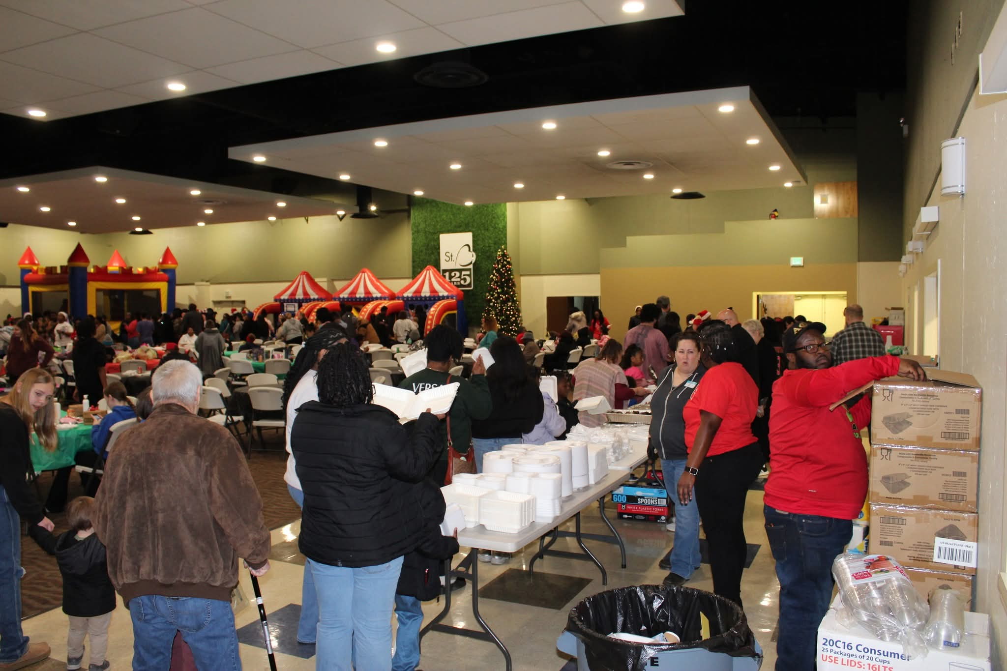 Crowded indoor event in a hall with long tables, plastic chairs, and inflatable red-and-yellow attractions in the background; a Christmas tree visible.