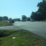Police cars with flashing lights blocking a busy highway near a grassy median and trees on a sunny day, with other vehicles stopped nearby.