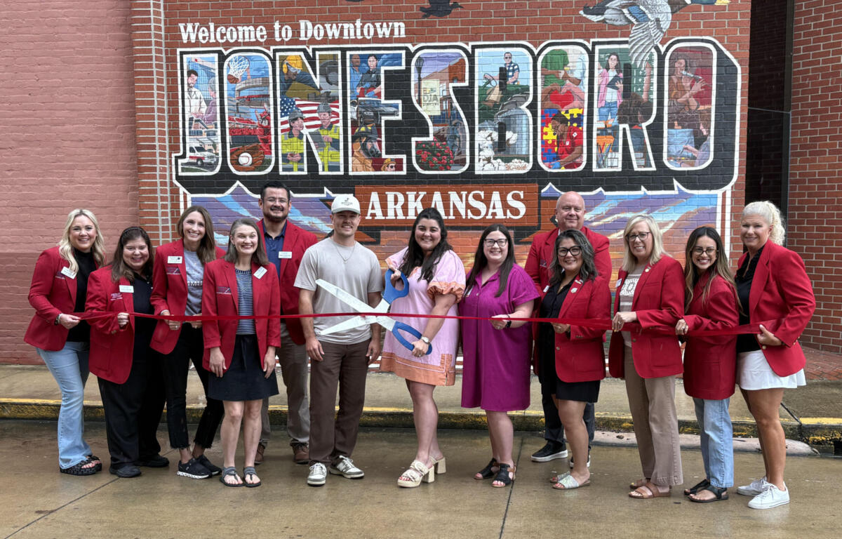 Group of people in red blazers at a ribbon-cutting in front of a large mural reading 'Welcome to Downtown Jonesboro, Arkansas'.