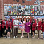 Group of people in red blazers at a ribbon-cutting in front of a large mural reading 'Welcome to Downtown Jonesboro, Arkansas'.