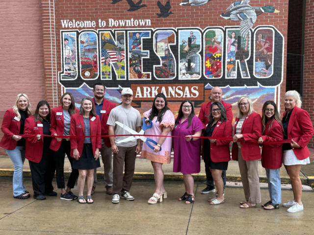 Group of people in red blazers at a ribbon-cutting in front of a large mural reading 'Welcome to Downtown Jonesboro, Arkansas'.