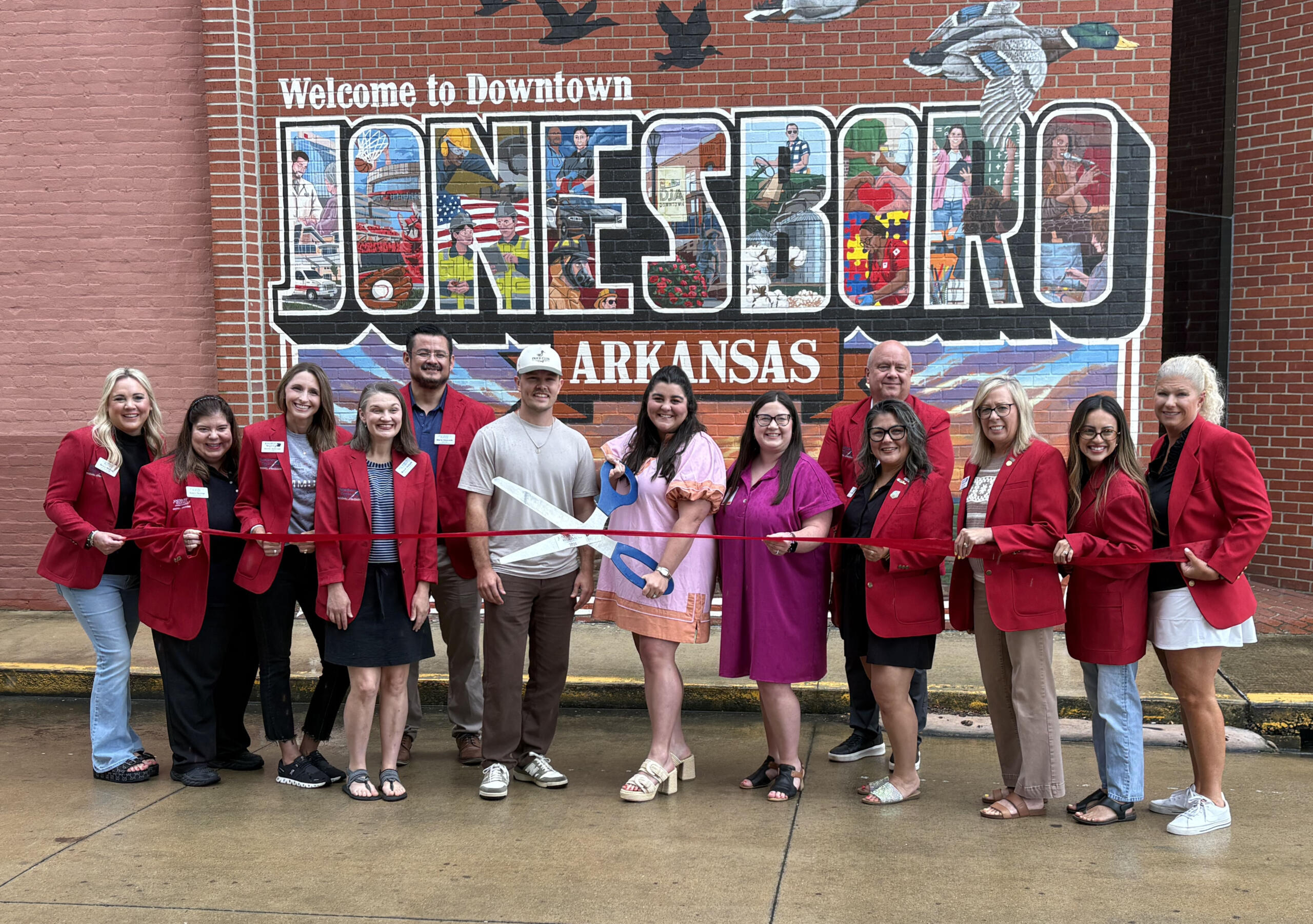 Group of people in red blazers at a ribbon-cutting in front of a large mural reading 'Welcome to Downtown Jonesboro, Arkansas'.