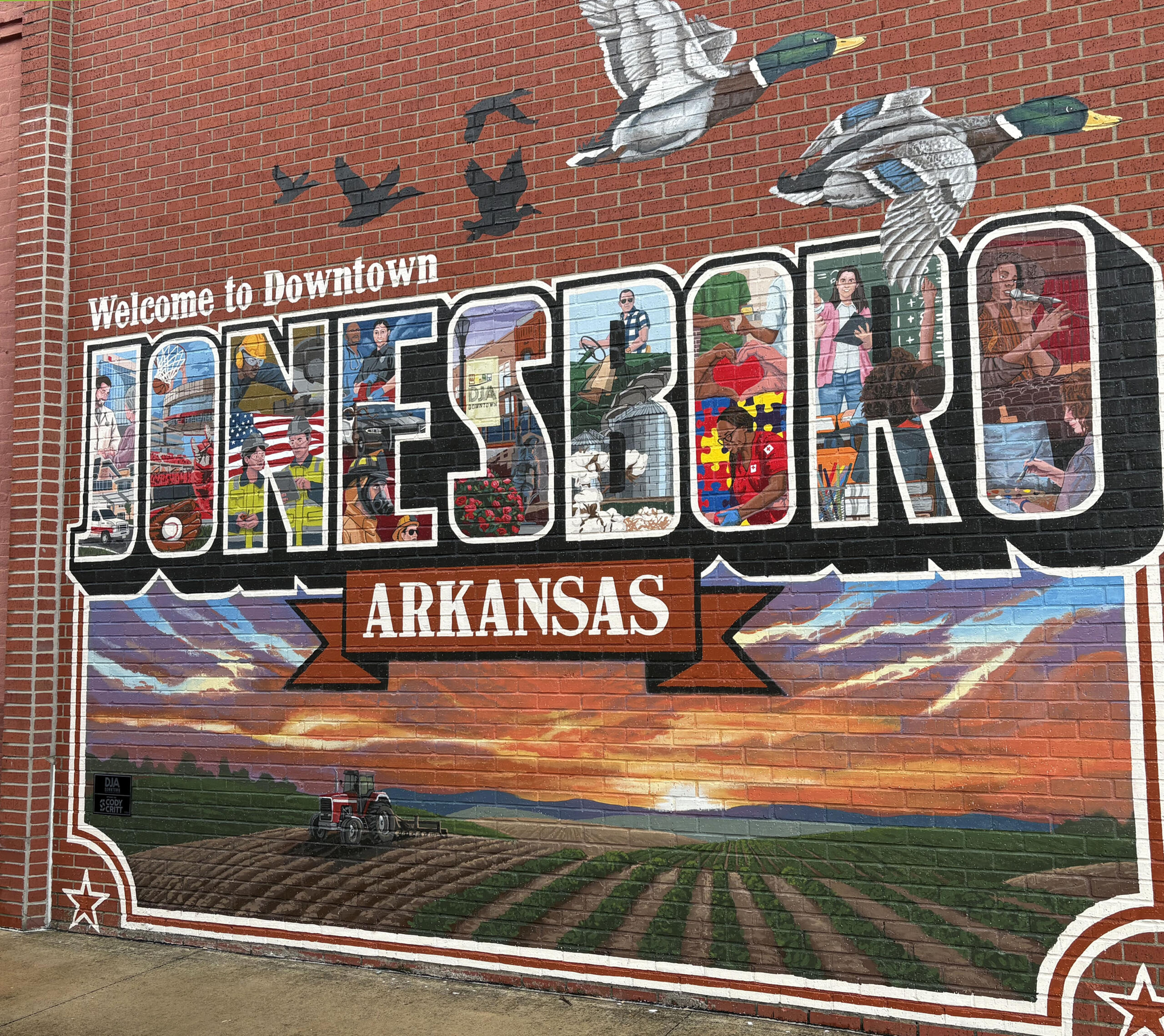 Mural on brick wall reading "Welcome to Downtown" with colorful windows and people depicted inside each letter, Arkansas imagery below.