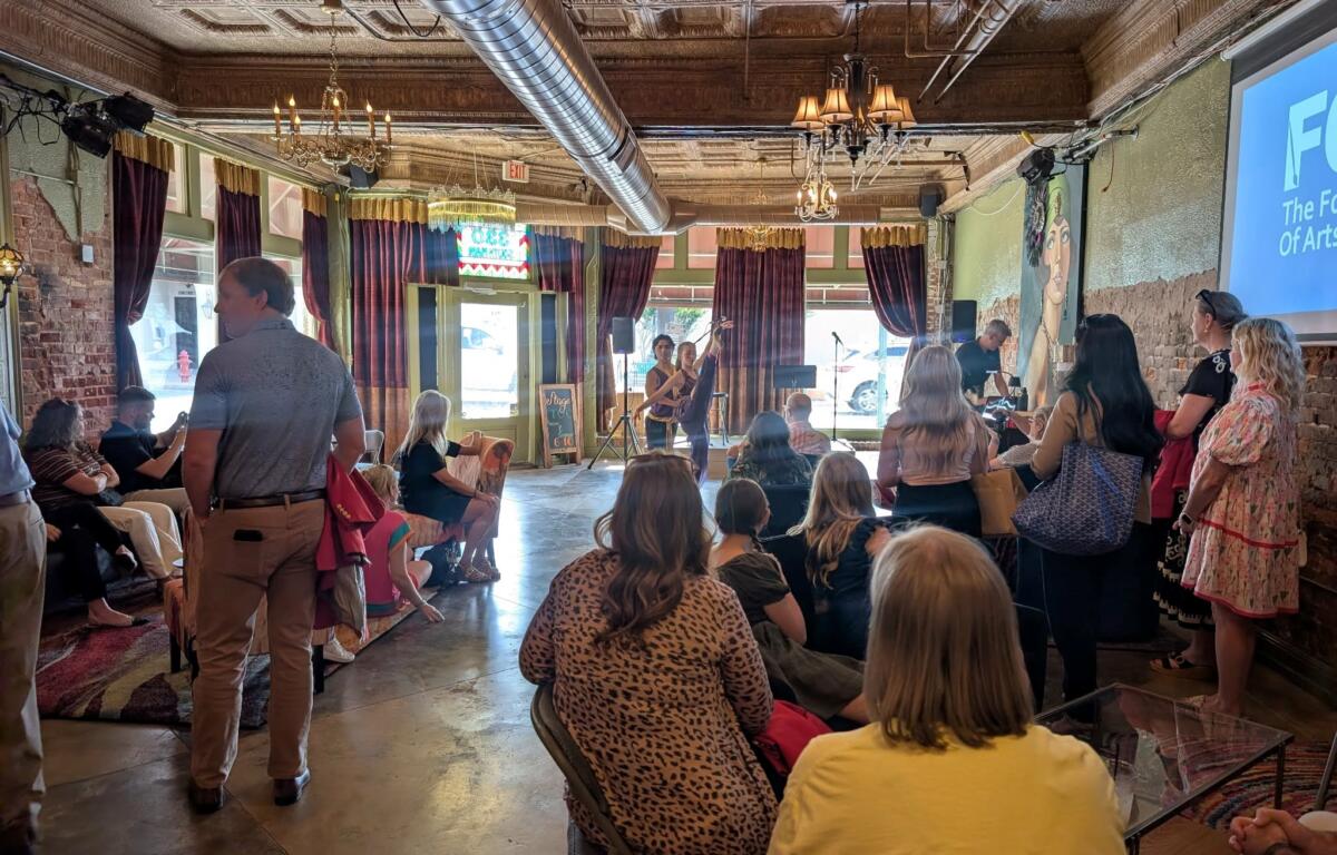 Audience seated and standing in a rustic venue watching a live performance on a small stage with a projector screen to the right.