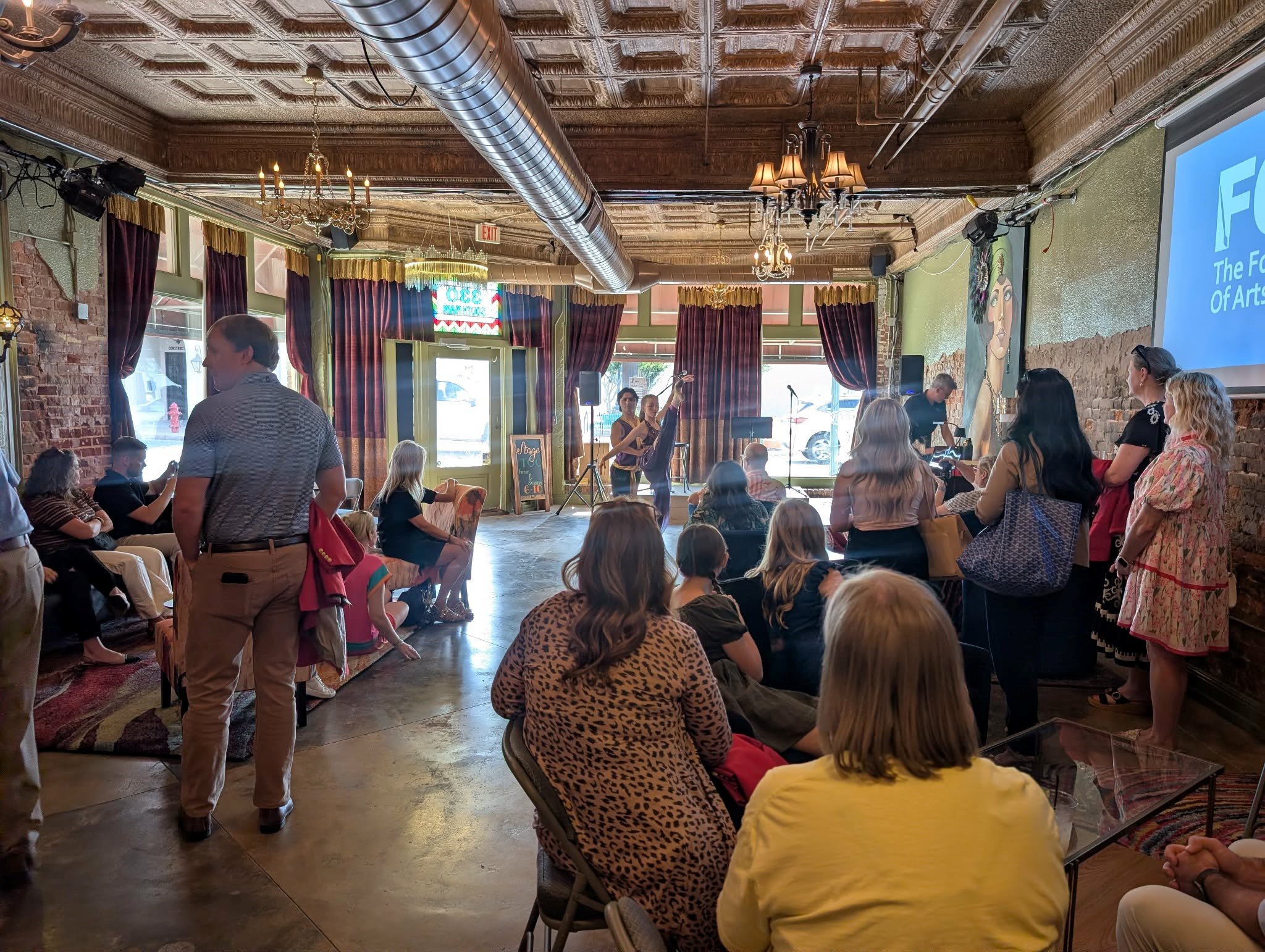 Audience seated and standing in a rustic venue watching a live performance on a small stage with a projector screen to the right.