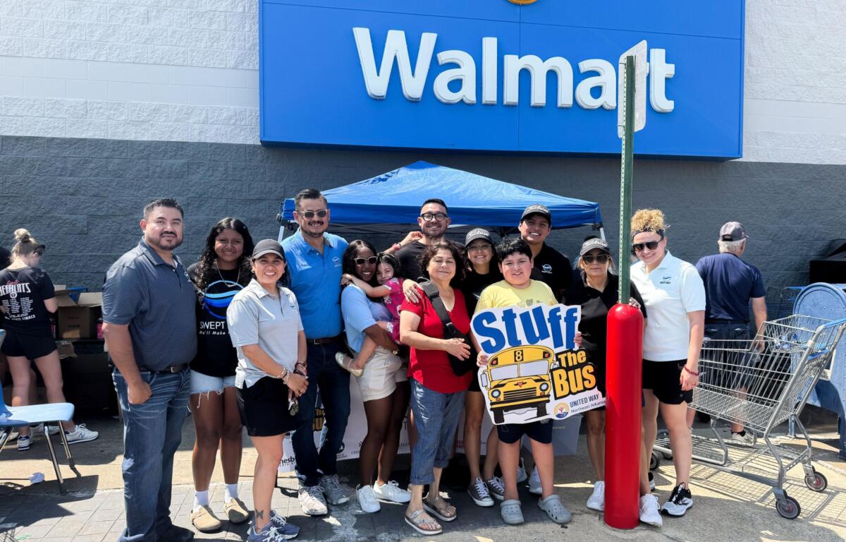 Group of diverse people posing for a photo outside a Walmart store under a blue sign, some smiles visible and a 'Stuff the Bus' poster nearby
