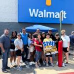 Group of diverse people posing for a photo outside a Walmart store under a blue sign, some smiles visible and a 'Stuff the Bus' poster nearby
