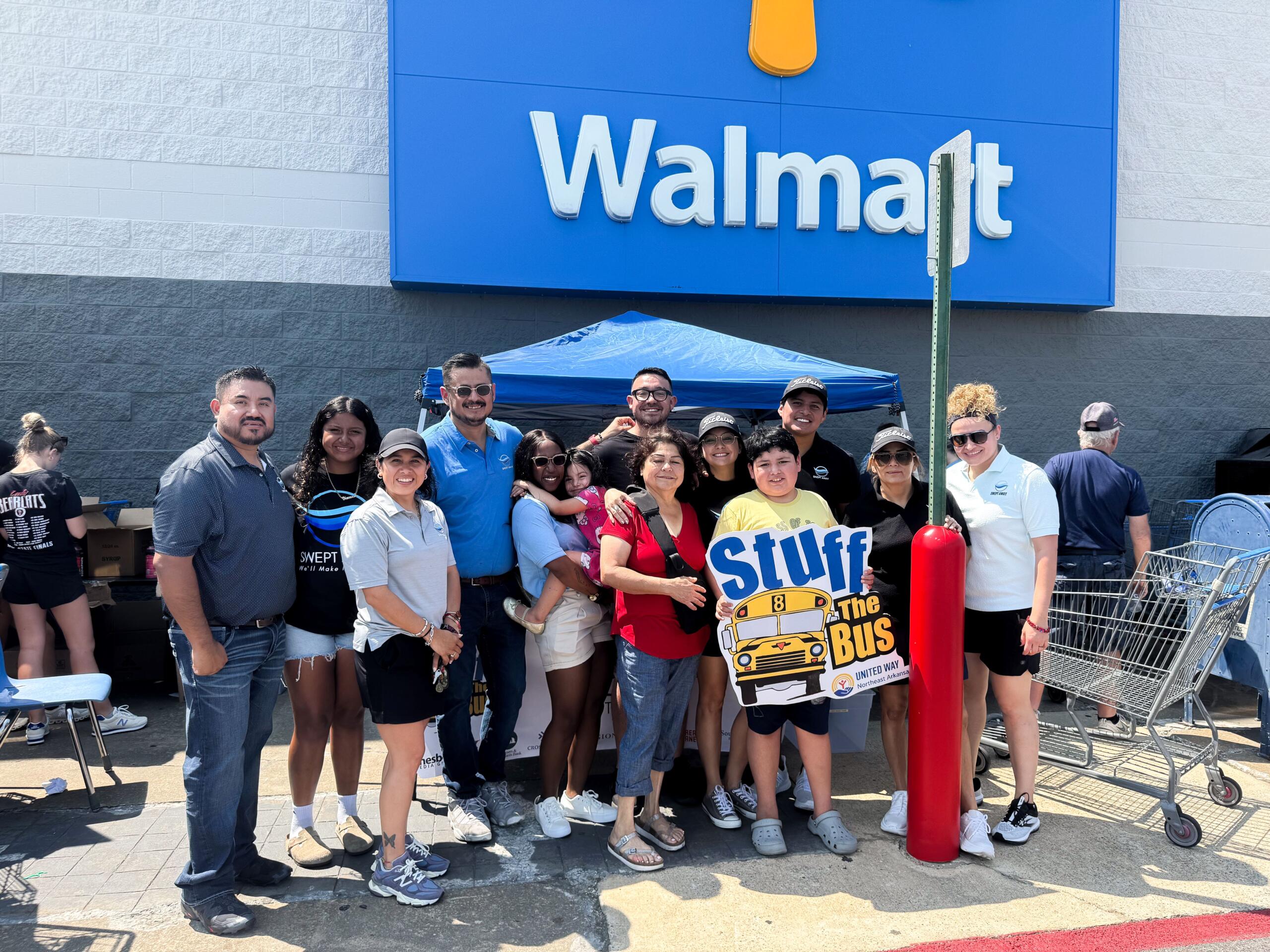 Group of diverse people posing for a photo outside a Walmart store under a blue sign, some smiles visible and a 'Stuff the Bus' poster nearby