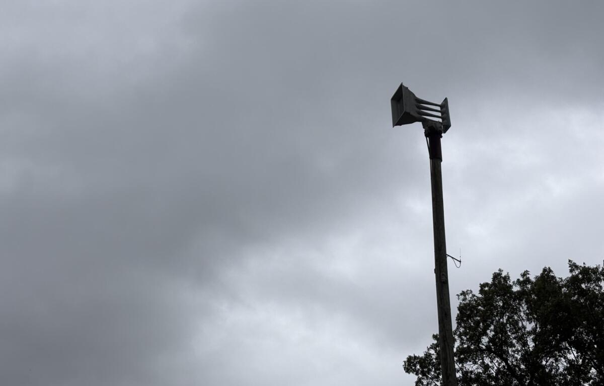 Tall utility pole with a multi-nozzle siren head extending horizontally, set against a gray, cloudy sky; trees visible on the right edge.