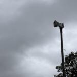 Tall utility pole with a multi-nozzle siren head extending horizontally, set against a gray, cloudy sky; trees visible on the right edge.