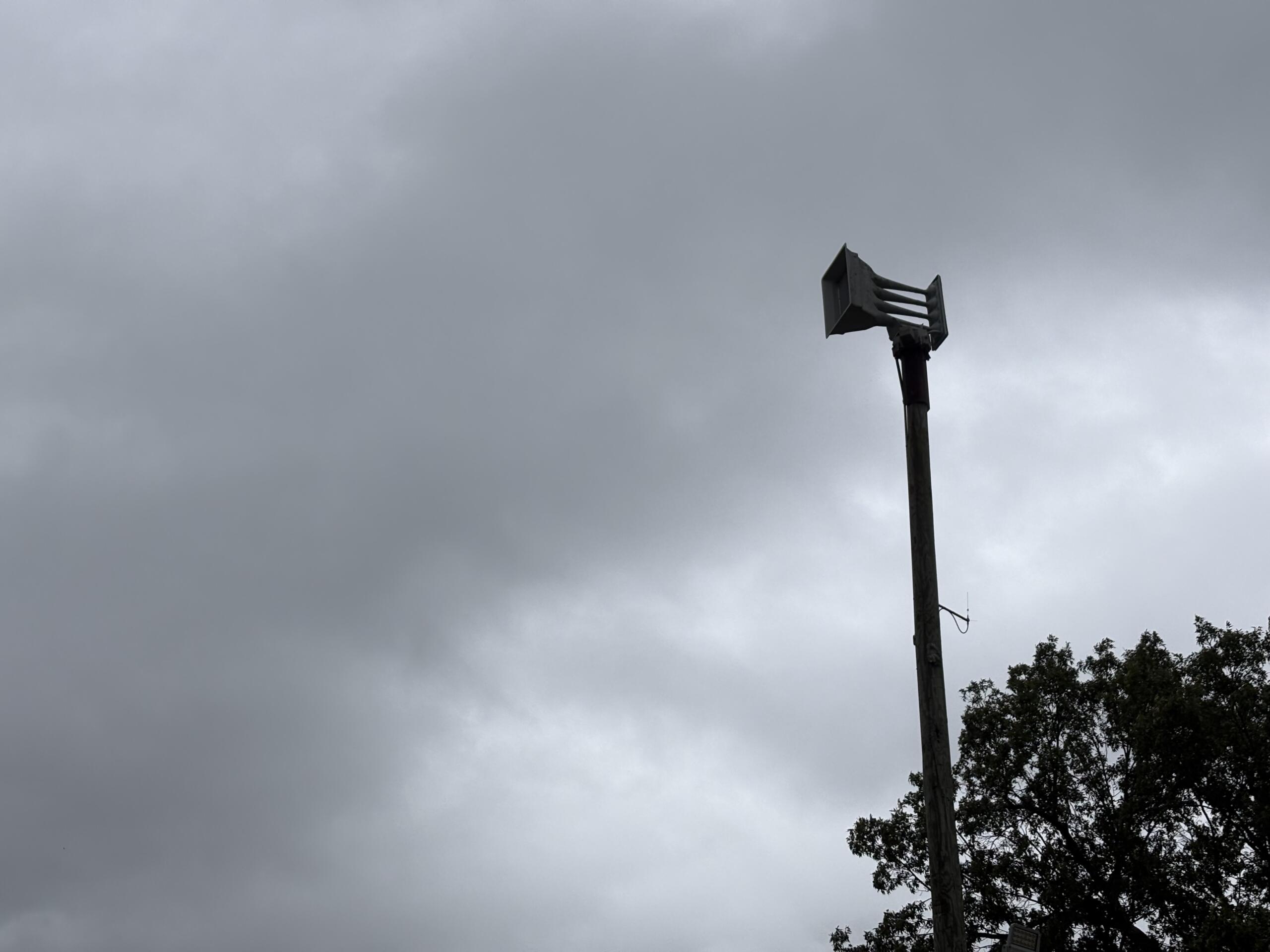 Tall utility pole with a multi-nozzle siren head extending horizontally, set against a gray, cloudy sky; trees visible on the right edge.